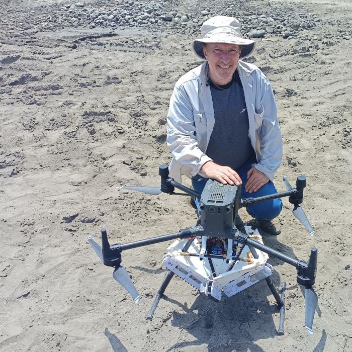 A man crouches next to a drone on land covered in gray sand or silt.  