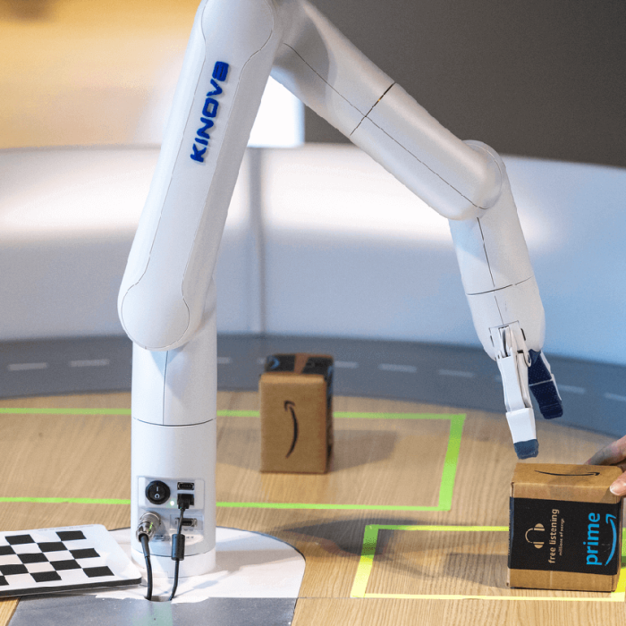 On a wooden table, a robotic arm reaches to pick up a small cardboard box being placed down by a human hand