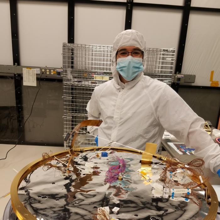 Technician stands wearing a white protective suit beside a circular piece of a spacecraft in-process
