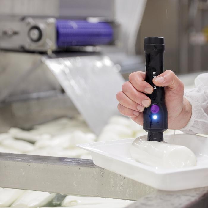 Close-up of a man’s hand holding a MicroNIR-branded device that is pressing against a white gelatinous substance in the shape of a cylinder in a tray