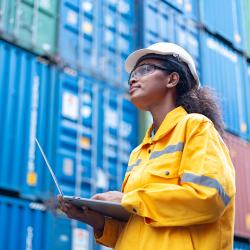 A woman holds a laptop in front of stacked shipping containers