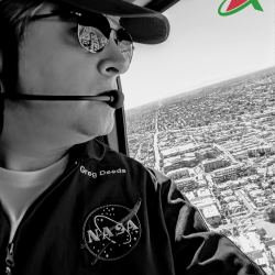 A black and white photo of a man wearing a shirt emblazoned with the NASA emblem looking out of the window of a helicopter at the city below.