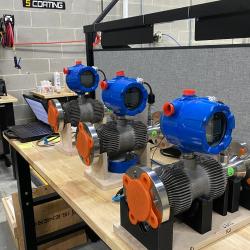 On a wooden table are three blue and orange machines next to a laptop computer