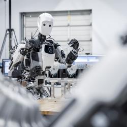 A white and black, humanoid robot holds a blade or machine part in a factory setting. 