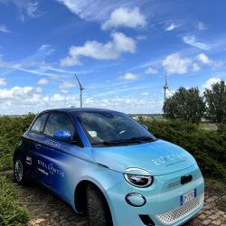 A blue compact car covered in decals advertising Electra and the company’s partners is parked on a brick road. In the background are trees, bushes, and wind turbines.