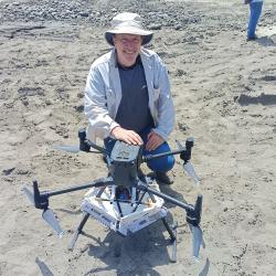 A man crouches next to a drone on land covered in gray sand or silt.  