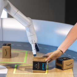 On a wooden table, a robotic arm reaches to pick up a small cardboard box being placed down by a human hand