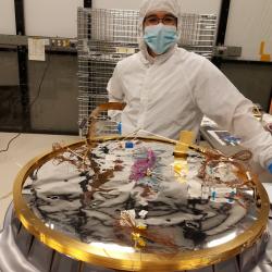 Technician stands wearing a white protective suit beside a circular piece of a spacecraft in-process