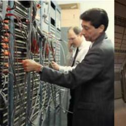 Two men plug and unplug wires attached to a wall of ports. Digital system covers the wall with no visible cables